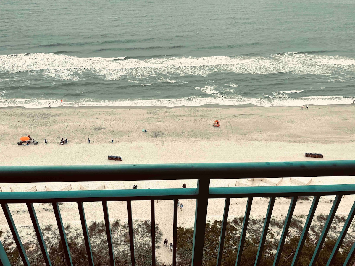 The image captures a serene ocean view from a private balcony. Gentle waves can be seen lapping against the sandy shore, where people are dotted along the beach. A lifeguard stand is visible in the distance, adding to the coastal atmosphere.