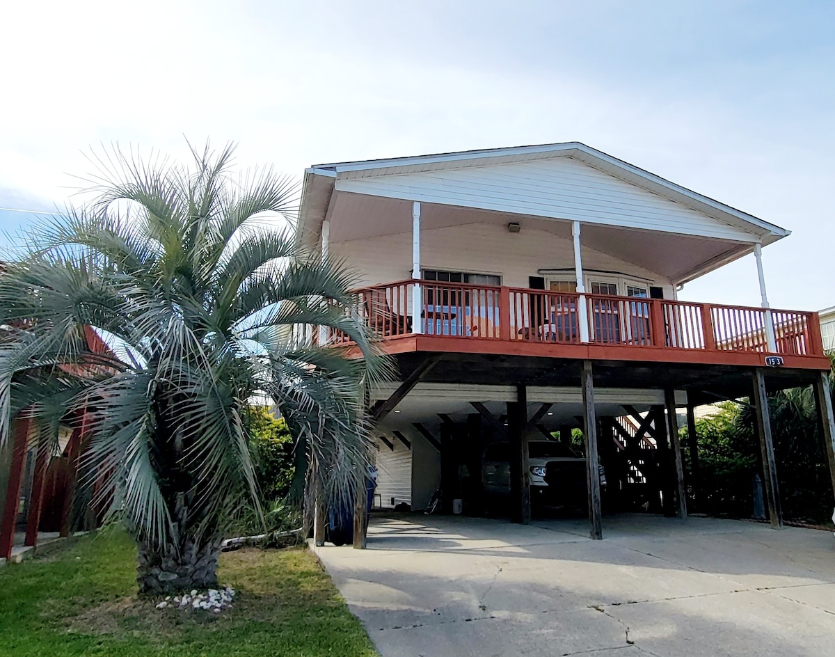 The exterior of the home features a raised structure with a spacious deck supported by wooden posts. Lush palm trees surround the area, and a concrete driveway provides access to the covered parking beneath the house.