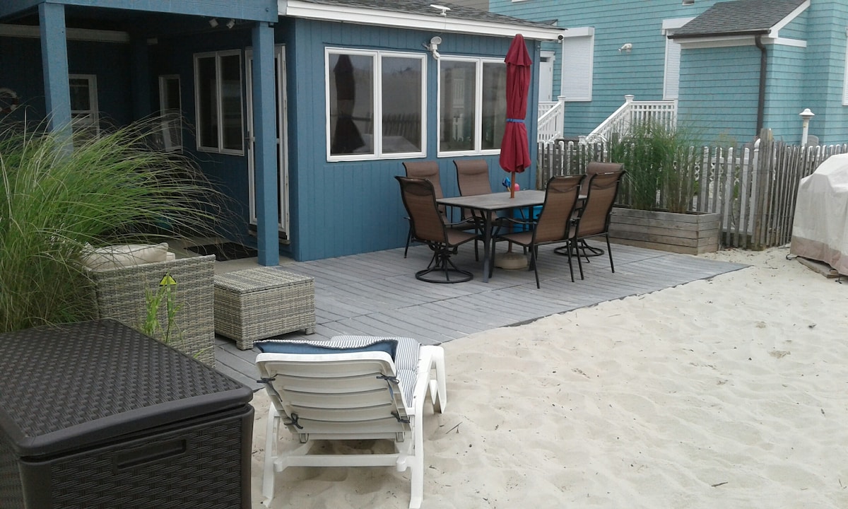 An outdoor dining area features a table with six sleek chairs and an umbrella, set against a backdrop of a blue bungalow. Nearby, a lounge chair and picnic seating are positioned on sandy ground, surrounded by decorative grass and wooden fencing.