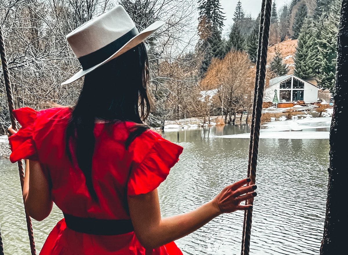 A woman stands on a swing, looking out towards a serene lake surrounded by trees. The landscape features a cabin in the distance, with muted tones of winter scenery reflected in the water's surface.