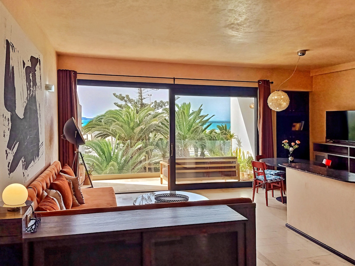 A spacious living area is illuminated by natural light from a large sliding glass door, showcasing views of palm trees and the sea. A comfortable sofa is positioned in front of a sleek TV unit, while a dining table sits nearby, enhancing the room's functionality.