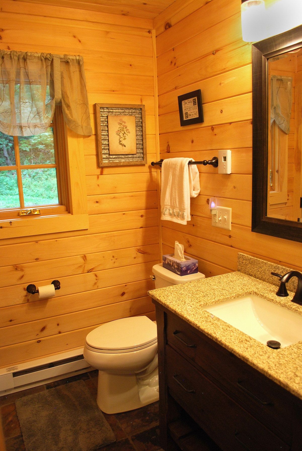 The bathroom features warm wooden walls that create a natural ambiance. A toilet is positioned beside a granite countertop sink with a large mirror above. Soft lighting is provided by a wall fixture, while a window allows natural light to enter. A towel hangs neatly on a rod.