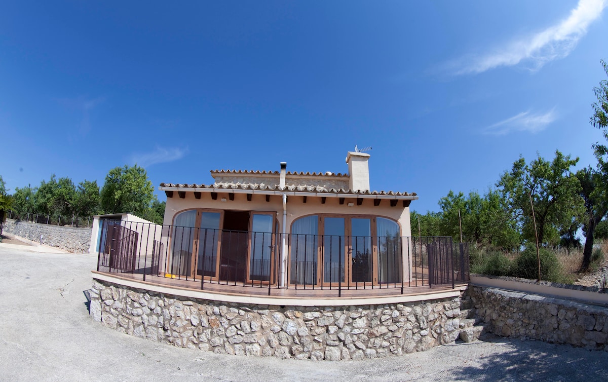 A spacious villa is positioned on elevated ground, featuring a terrace with a stone wall. Large windows provide natural light to the interior, while a pathway leads toward the entrance. Lush greenery surrounds the villa, contributing to a serene rural setting.