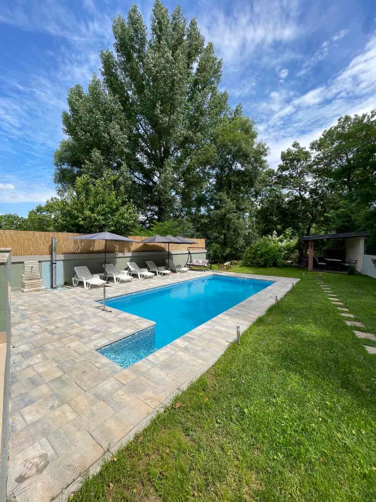 A serene swimming pool is surrounded by well-maintained greenery and paved stone tiles. Several lounge chairs with canopies provide comfortable seating, while a shaded area is visible nearby, enhancing the outdoor relaxation space.