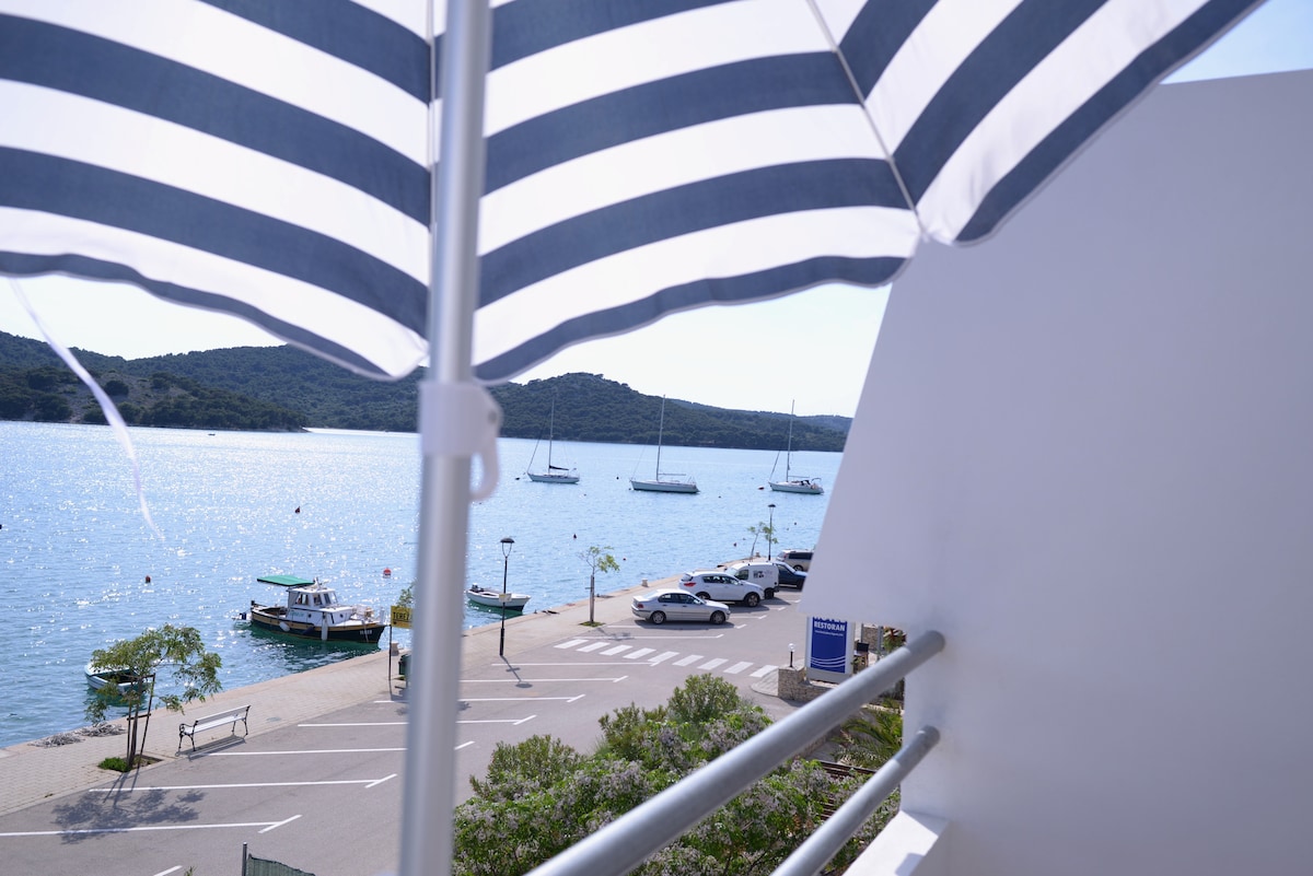 A seaview balcony presents a view of a tranquil harbor, featuring several moored sailboats and small fishing vessels. A white and navy striped sunshade is overhead, gently fluttering in the breeze, while the surrounding area includes lined parking spaces and lush greenery near the waterfront.