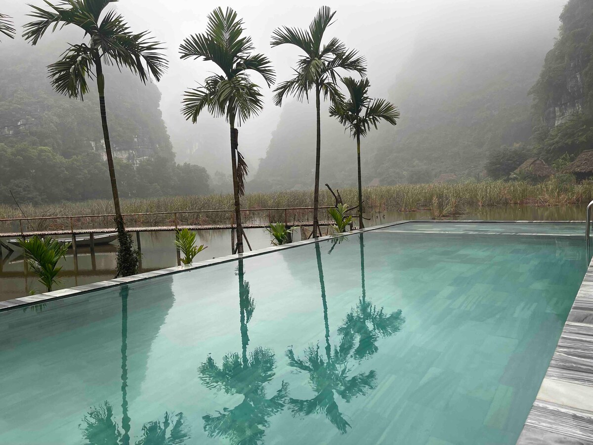 An expansive swimming pool reflects the surrounding misty landscape, featuring tall palm trees framing the view. Soft ripples in the water highlight the natural beauty of the high mountains in the background, creating a serene and tranquil atmosphere.