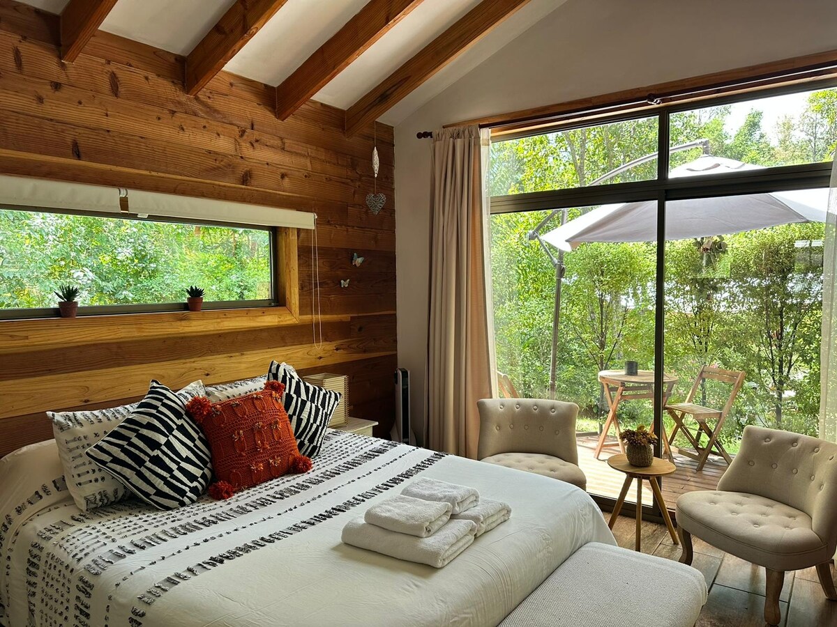 A cozy bedroom features wooden walls that create a warm ambiance. A spacious bed is adorned with decorative pillows. Natural light filters through large glass doors, which open to a private terrace with two outdoor chairs and a table surrounded by lush greenery.