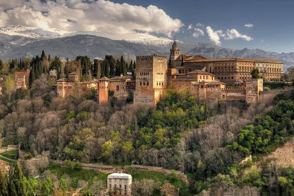 A panoramic view displays the Alhambra fortress, nestled against a backdrop of mountains. The architectural details of the fortress are visible, framed by surrounding trees and greenery. The sky features a mix of clouds and sunlight, enhancing the serene landscape.
