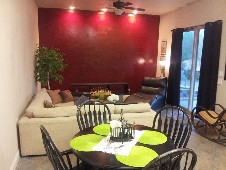 An inviting living space features a sectional sofa against a rich red accent wall, with a small dining table set for four. A rocking chair and a potted plant add to the cozy atmosphere, while large windows allow natural light to enter the room.