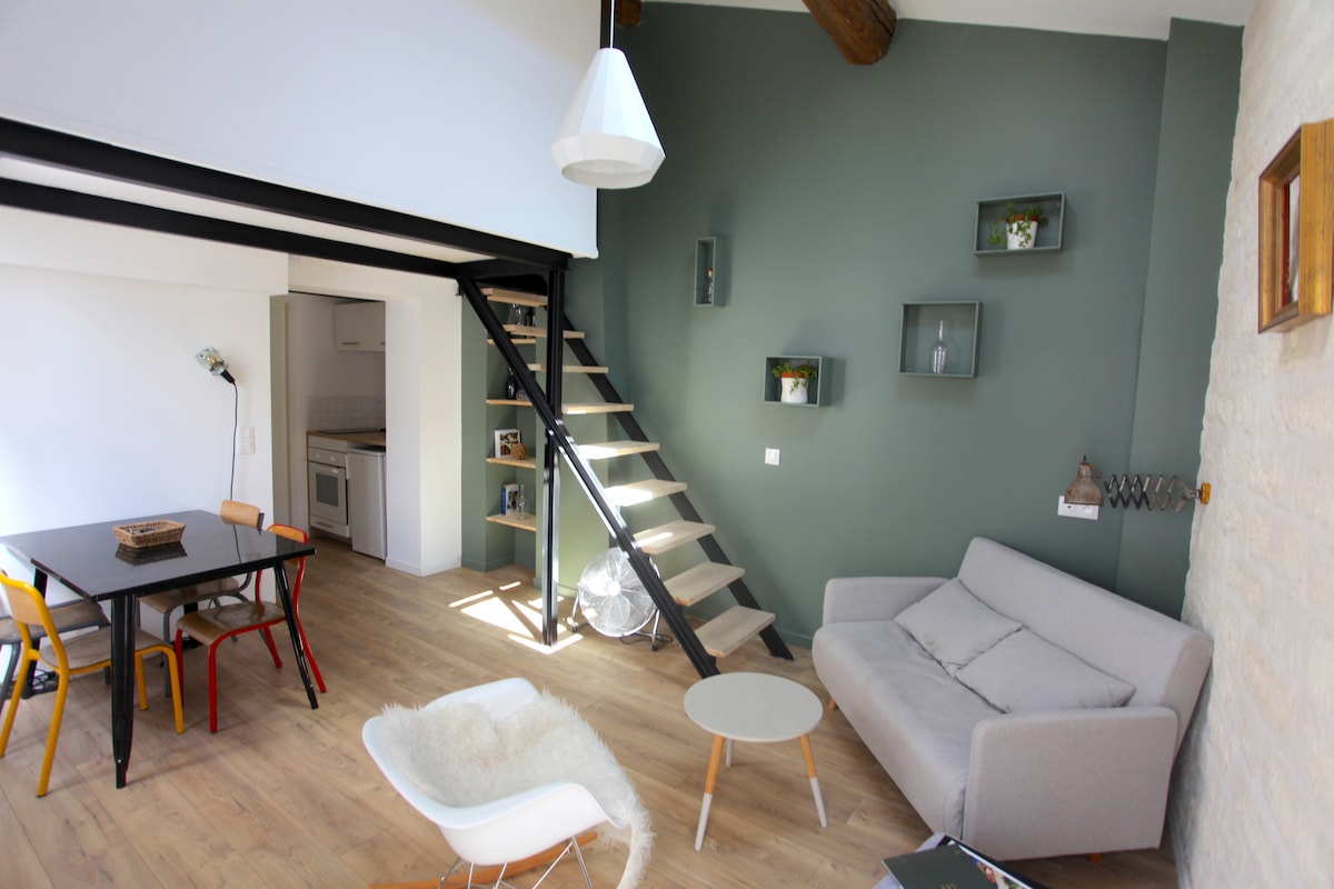 A cozy living area features a compact, modern sofa and a small round coffee table. A wooden staircase leads to the upper level, accentuated by green walls and minimalist shelves. A dining table with colorful chairs complements the light hardwood flooring.