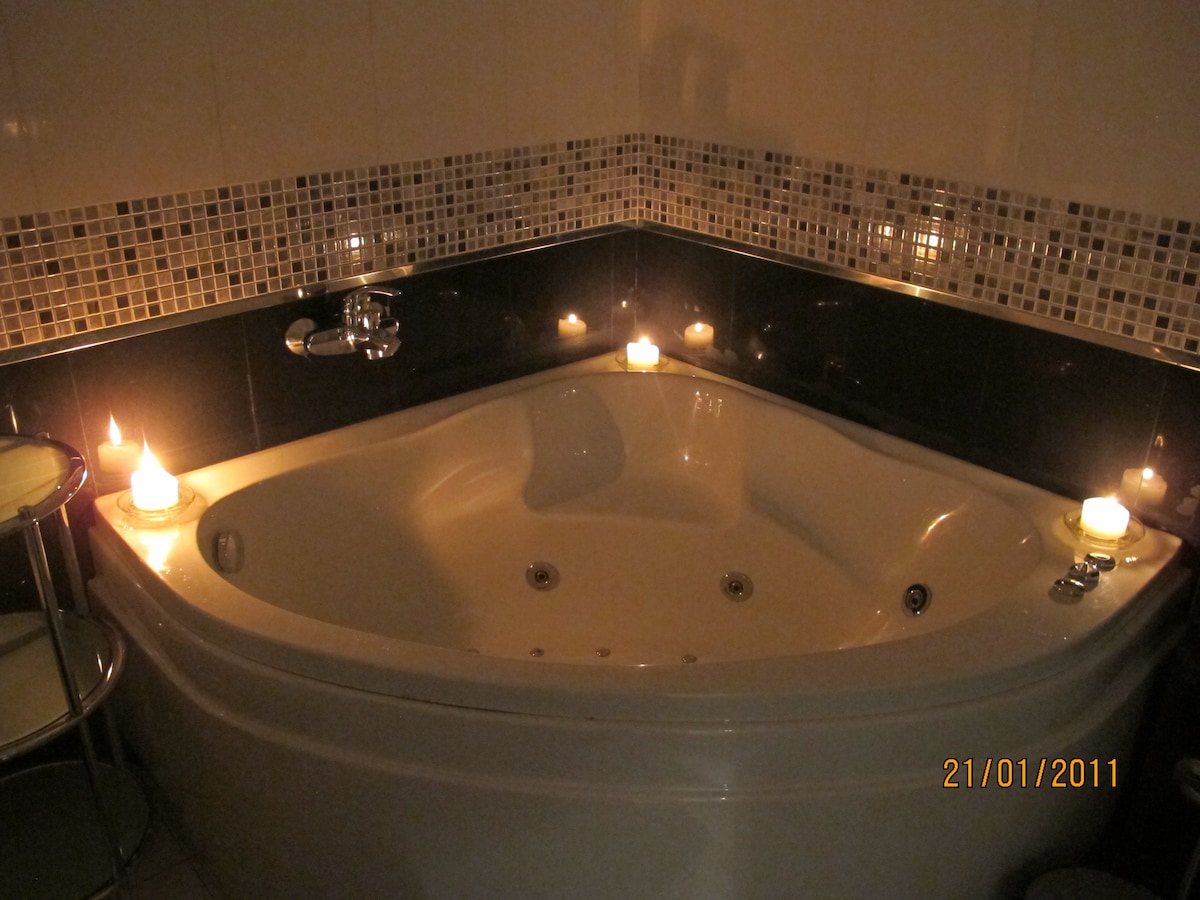 A spacious whirlpool bath is surrounded by decorative tiles and softly lit by candles placed on the edges. The bath features a sleek design, emphasizing comfort and relaxation, while the calming glow of the candles creates a soothing ambiance.