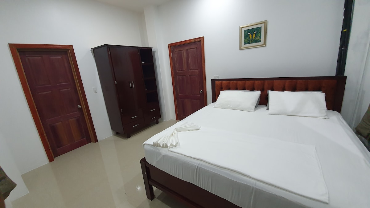 A spacious bedroom is furnished with a modern bed topped with white linens and pillows. A dark wooden wardrobe stands against one wall, while two closed doors are visible in the background. Soft lighting illuminates the room, enhancing the neutral color scheme.
