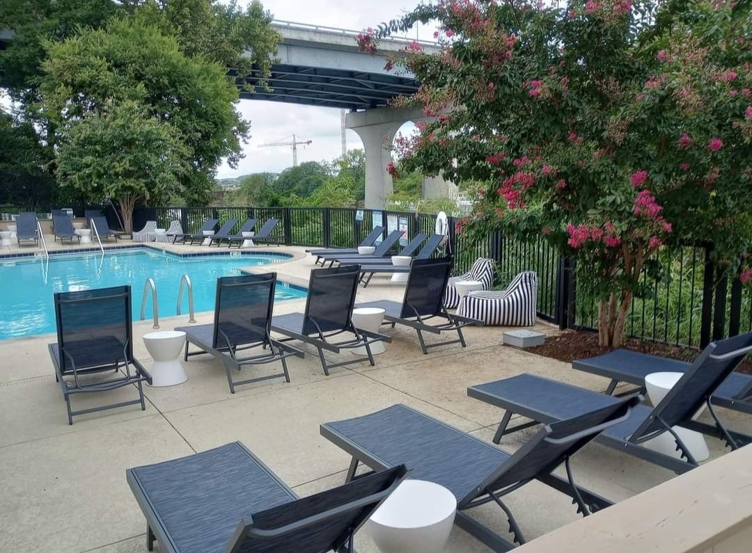 An outdoor pool area is surrounded by lounge chairs in a modern design. A mix of sturdy loungers and striped bean bags sit beside the clear blue water. Lush greenery and flowering plants create a serene ambiance, while a bridge is visible in the background.