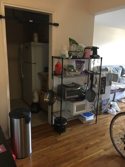 standing in the doorway, kitchen on the left. Food rack has microwave and toaster oven. Bed and couch can be seen in the background on the right.