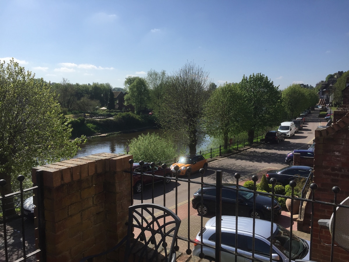 A clear view of the river Severn is presented, framed by lush green trees lining the banks. Cars are parked along the quiet road, and a railing can be seen in the foreground, enhancing the sense of openness in the inviting atmosphere.