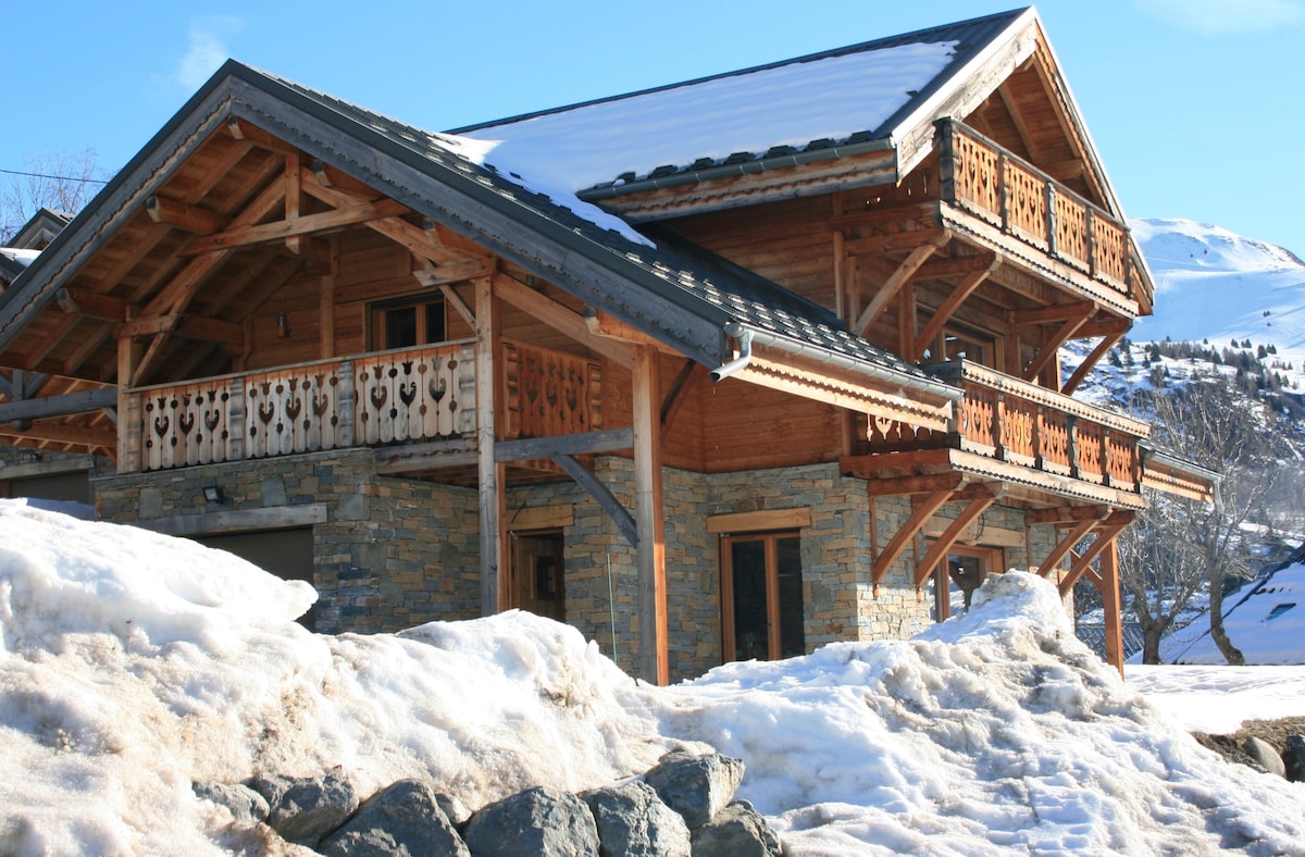 The chalet features a charming alpine design with a combination of natural stone and wooden elements. A prominent balcony with decorative railings is visible on the upper level, while a layer of fresh snow covers the ground and rooftops, creating a serene winter setting.