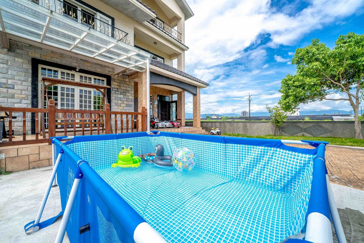 A small above-ground pool, brightly bordered in blue, is invitingly filled with clear water. Colorful pool accessories, including a frog and a beach ball, are floating on the surface. The setting features a spacious outdoor area with a stone facade and greenery in the background.