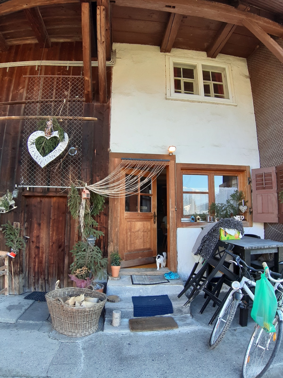 The entrance to a rustic farmhouse is framed by wooden features, displaying a heart-shaped decoration and hanging plants. A welcoming door opens into the home, while potted plants and a bicycle are positioned nearby, enhancing the inviting exterior ambiance.