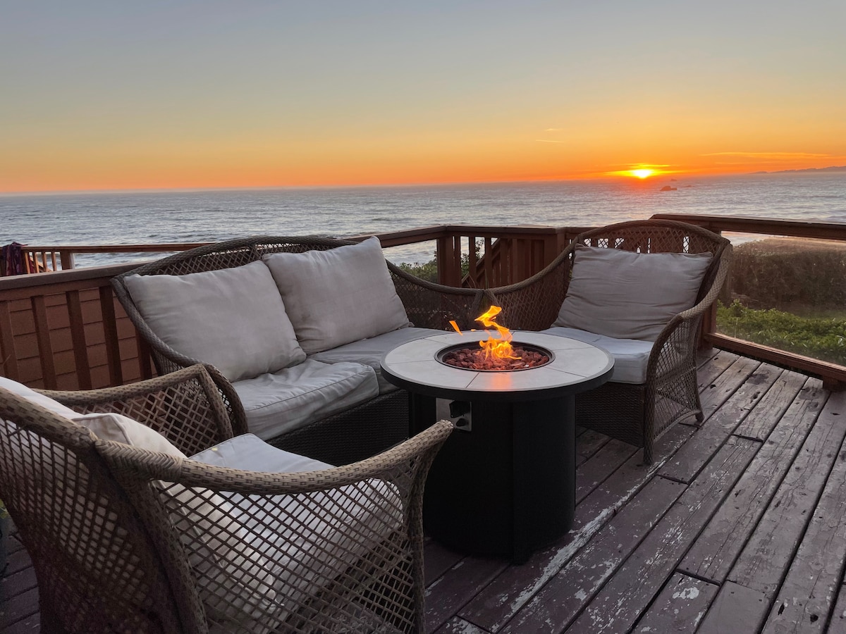 A comfortable seating area is arranged on a wooden deck, featuring two woven chairs and a round fire pit table. The scene is set against a captivating sunset over the ocean, with colors ranging from warm oranges to soft purples in the sky.
