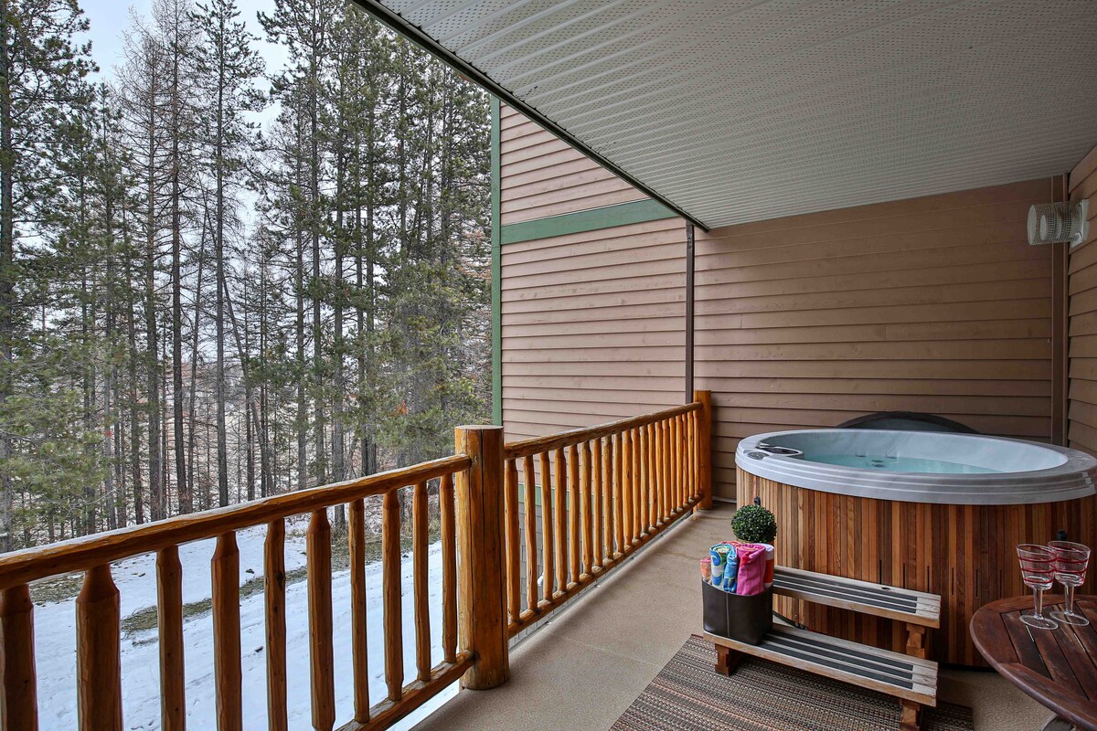 A private deck features a wooden railing and a cozy hot tub, surrounded by pine trees. A small table with two glasses and a decorative basket is positioned nearby, creating an inviting space to relax. The snowy landscape is visible beyond the deck.