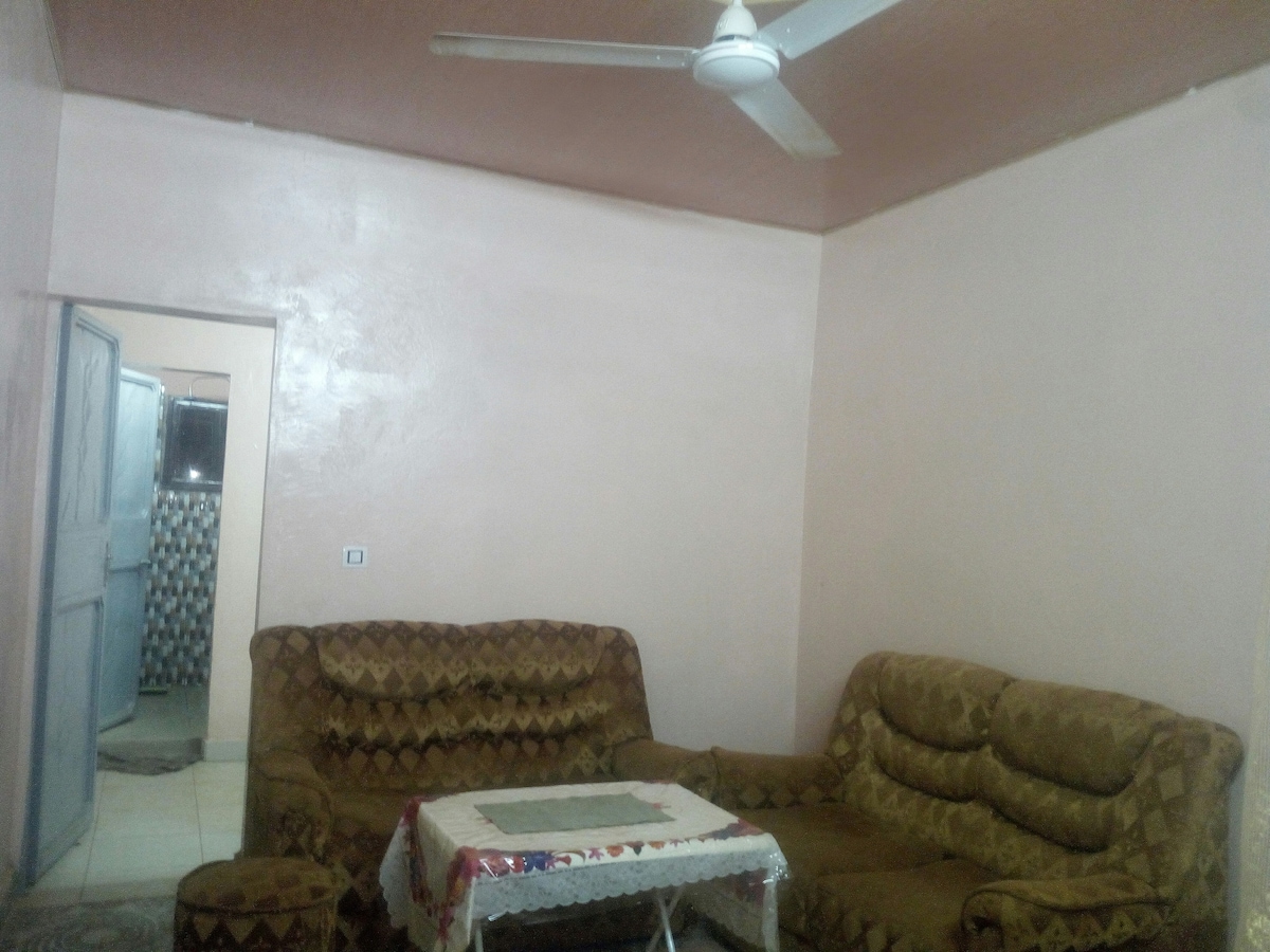A living area is presented with two patterned sofas arranged around a white, lace-covered table. A ceiling fan is visible above, and a doorway leads to another space. The walls are painted in a soft pink hue, creating a simple and functional environment.