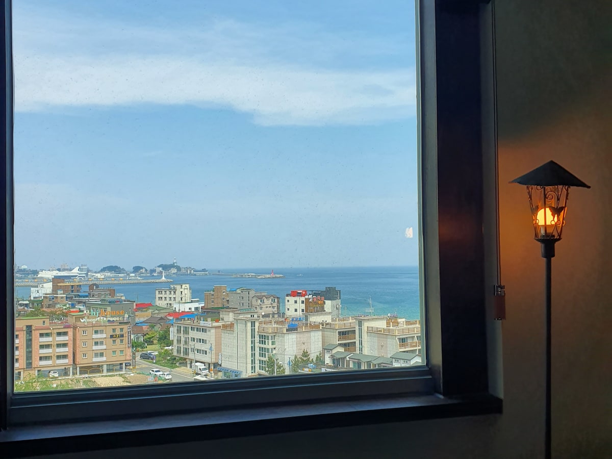 A view from the window captures the serene horizon where the sea meets the sky, with a mix of coastal buildings in the foreground. A softly glowing lamp adds a touch of warmth beside the window.