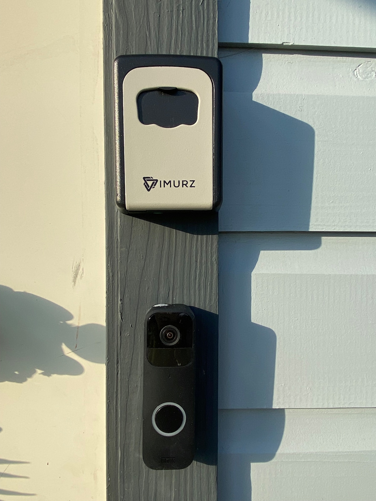 A doorbell camera and a secure lockbox are mounted on the exterior wall near the entrance. The camera features a circular button and is positioned below the lockbox, which has a modern design. The wall displays a soft color, complementing the entrance.