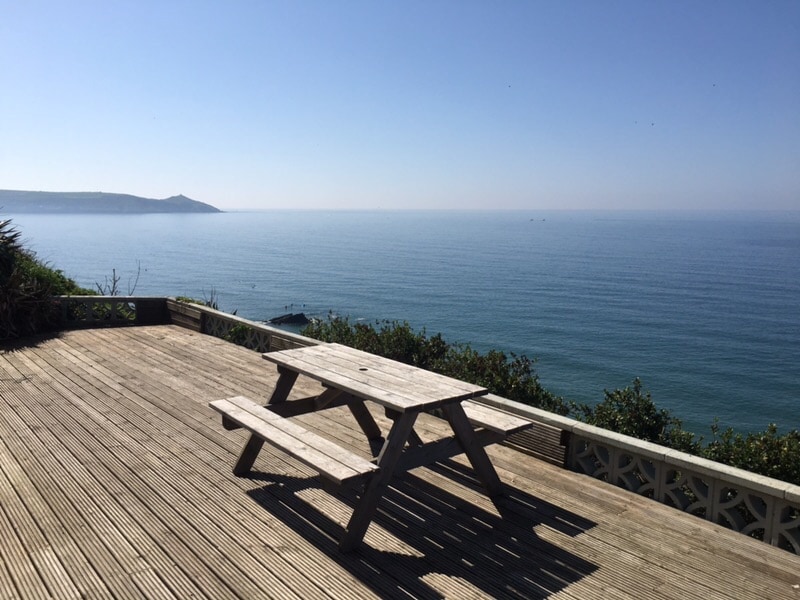 A spacious wooden decking area is visible, featuring a picnic table that invites relaxation. The unobstructed sea view stretches across the horizon, reflecting the bright blue sky, while the tranquil waters gently ripple in the distance.