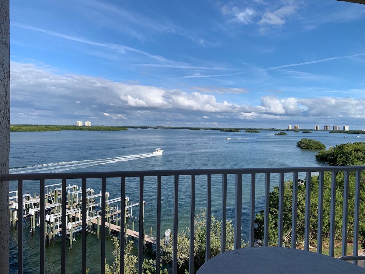 A panoramic view of Estero Bay is visible from the balcony, featuring calm waters and small islands. Boats can be seen moving along the water, while lush greenery and docks frame the scene. Fluffy clouds drift across a bright blue sky.