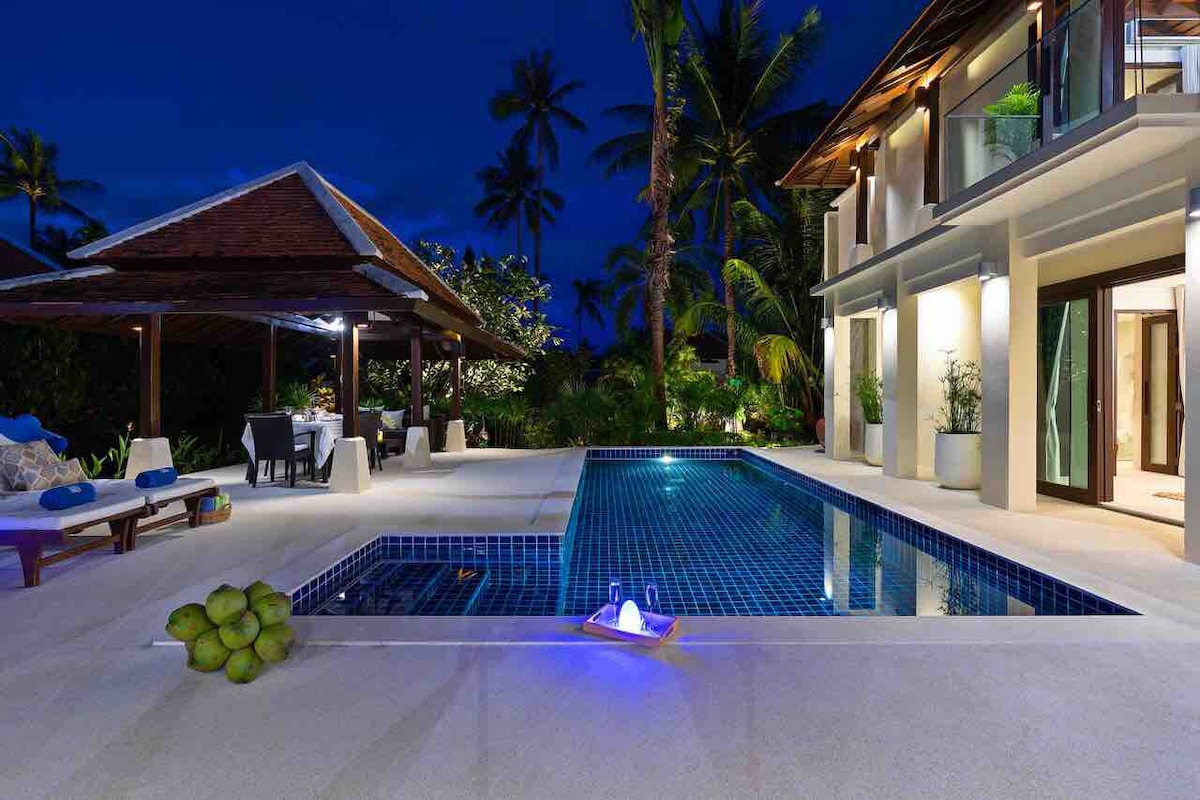 A tropical villa is showcased at night, featuring a private swimming pool with gentle blue lighting. Surrounding palm trees frame the space, and a covered seating area with dining options is visible nearby, providing a serene atmosphere for relaxation.