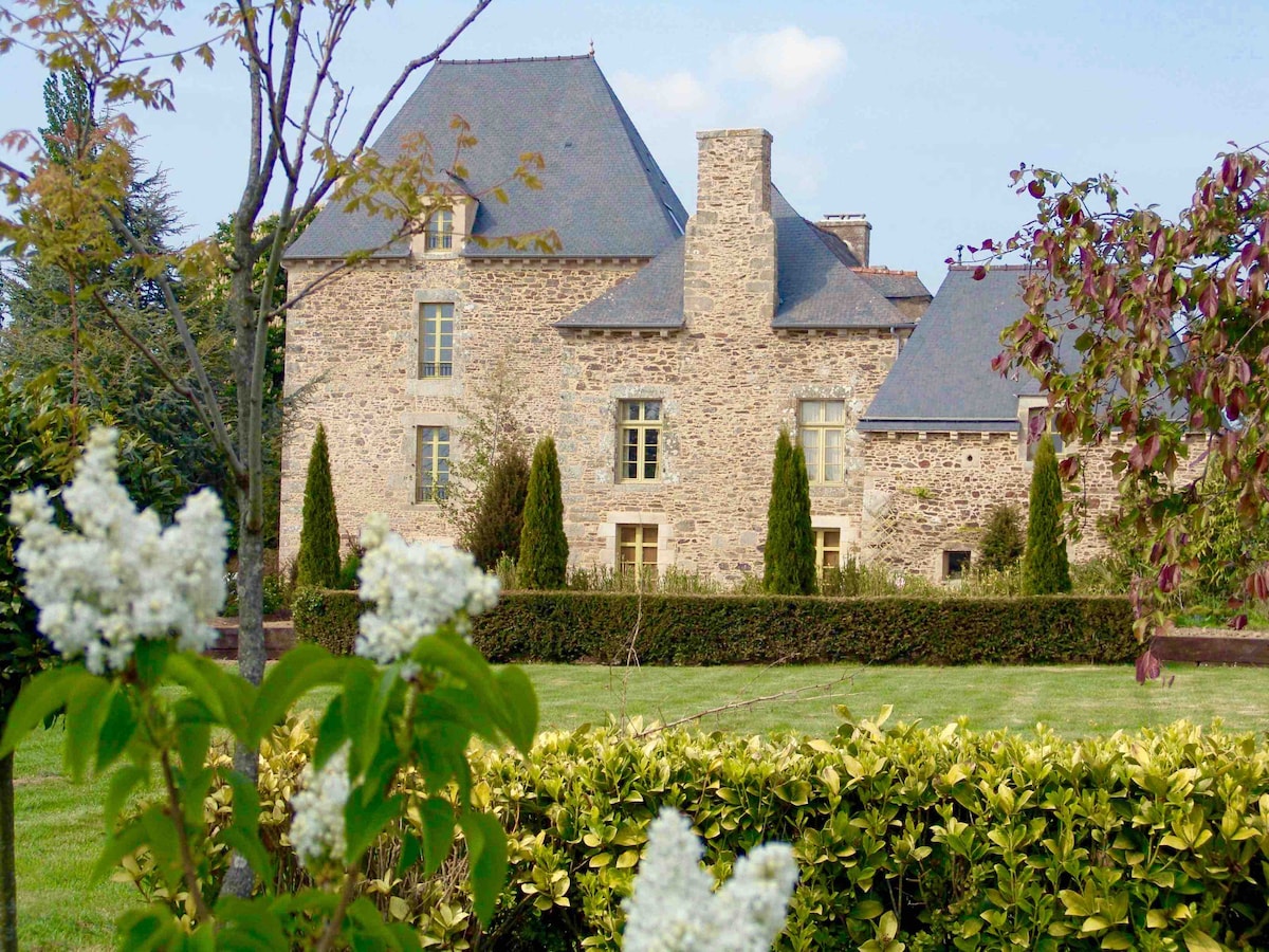 The historic manor is showcased with its stone facade and traditional architecture. Lush greenery surrounds the property, with neatly trimmed hedges and blooming flowers in the foreground, creating a serene and inviting outdoor space.