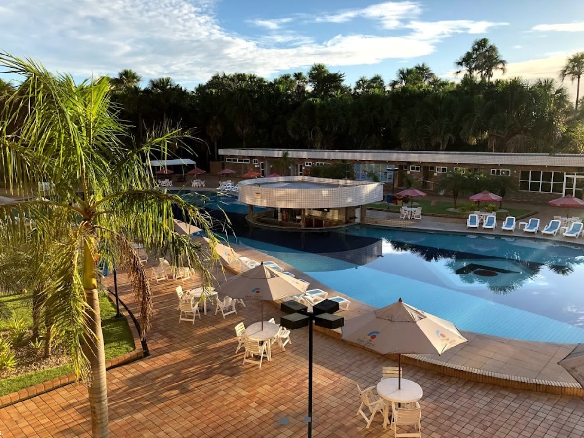 An outdoor pool area is surrounded by palm trees and lounge chairs. Umbrellas provide shade over tables, while the pool features a circular bar in the center. The serene environment is complemented by the green landscape visible in the background.