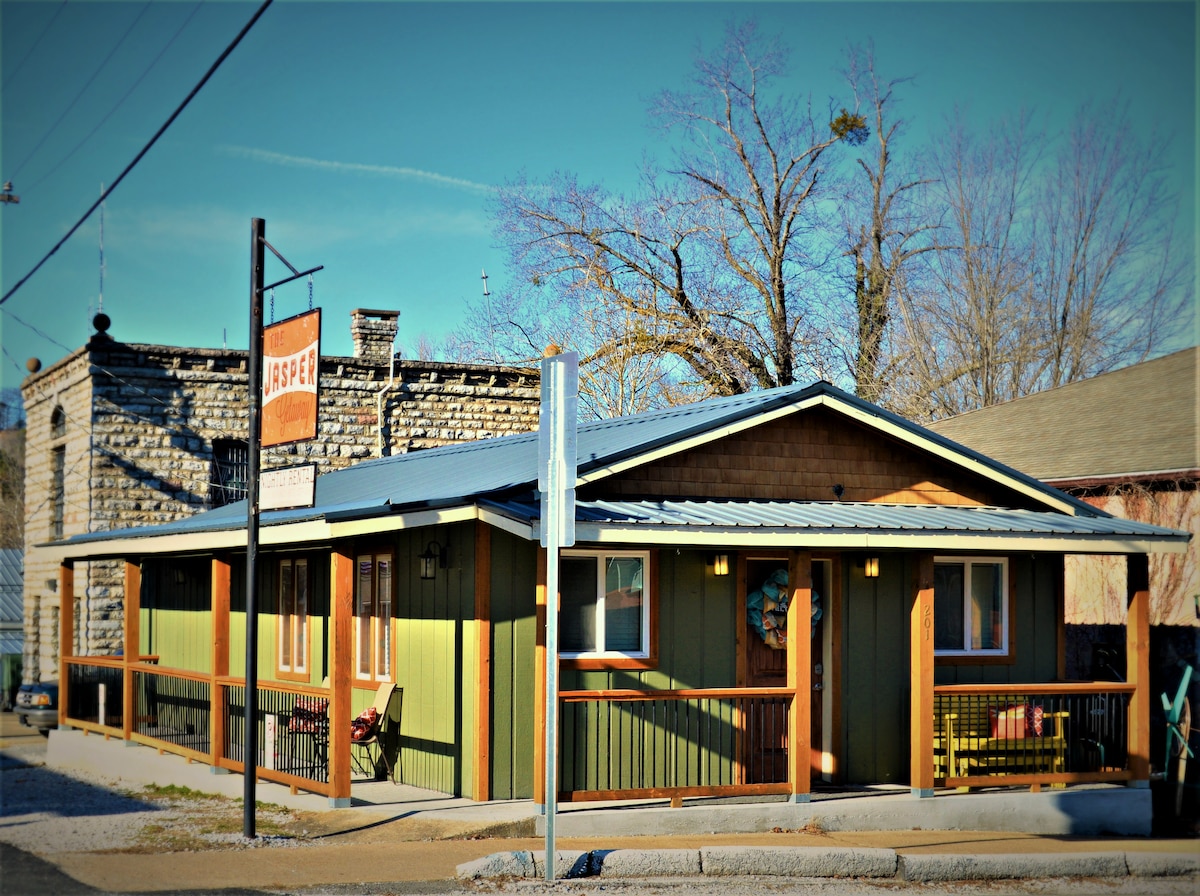 The exterior of the Jasper Getaway is captured, showcasing a modern cabin with green and wooden accents. A welcoming sign hangs above the entrance. The surrounding area features manicured landscaping and a clear sky, creating a charming atmosphere in downtown Jasper.