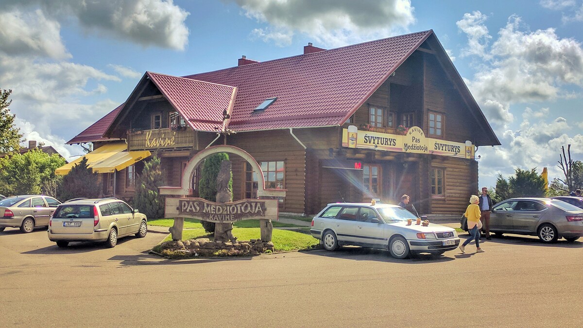 The exterior of the motel is showcased, featuring a charming wooden structure with a red roof. A welcoming sign displays the name 'Pas Medžiotoją.' Several parked cars are visible in the foreground, along with green landscaping and a clear blue sky in the background.
