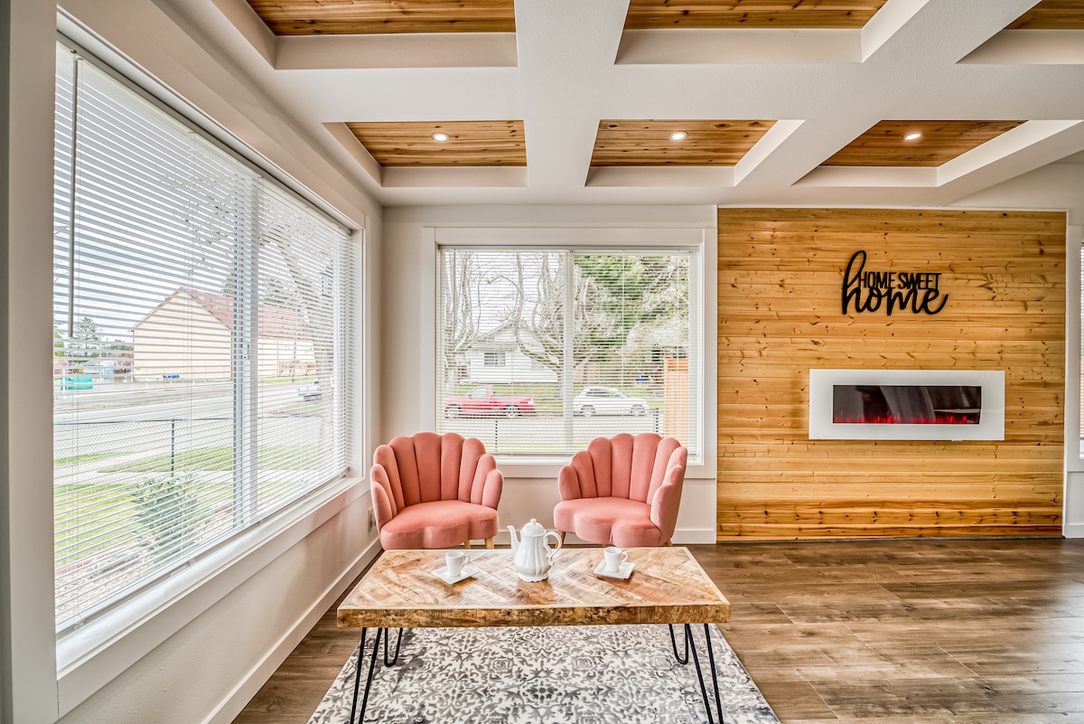 A bright seating area features two pink chairs positioned beside a low wooden table, set with a tea service. Large windows allow natural light to fill the space, and a wooden accent wall adds warmth. A decorative sign displaying 'Home Sweet Home' complements the decor.