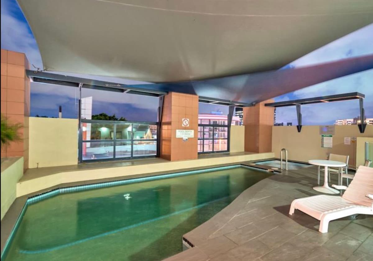 An outdoor swimming pool is visible, framed by a covered area that offers shade. A few lounge chairs are placed nearby, and a small table is located at one end. The surroundings are enhanced by soft evening lighting, creating a calming environment.