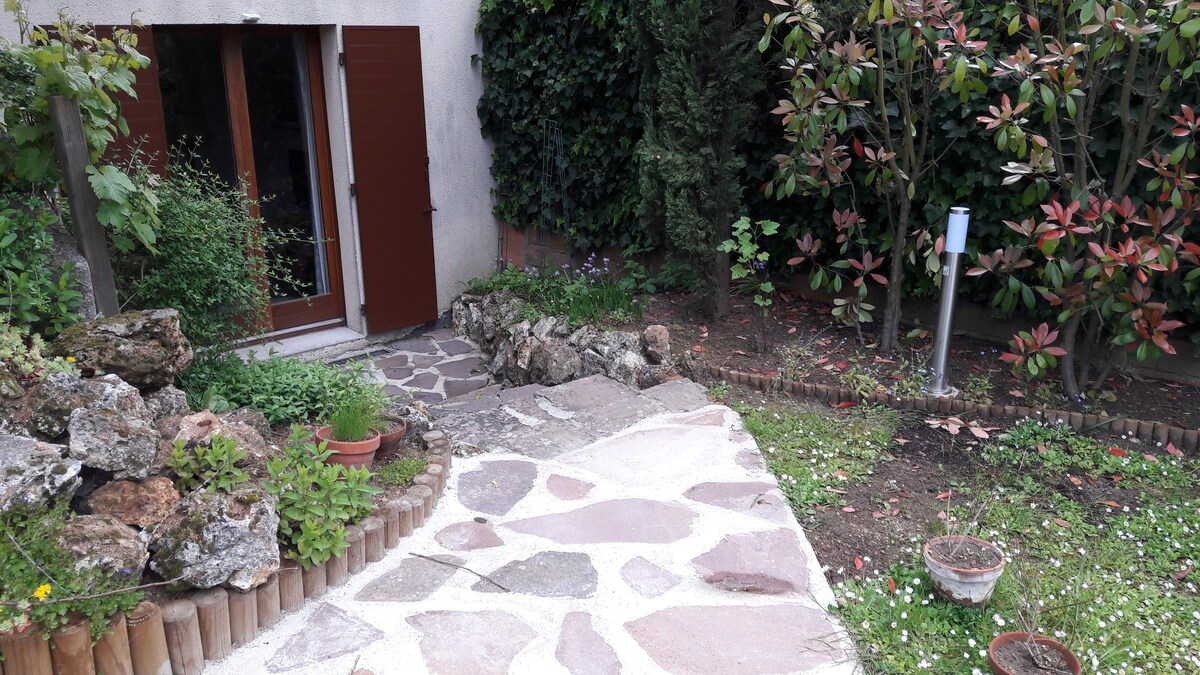 A garden pathway leads to a set of wooden doors, surrounded by lush greenery and flowering plants. Natural stone pavers create a textured surface, while potted plants are arranged along the edges. The setting provides a serene outdoor space, ideal for relaxation.
