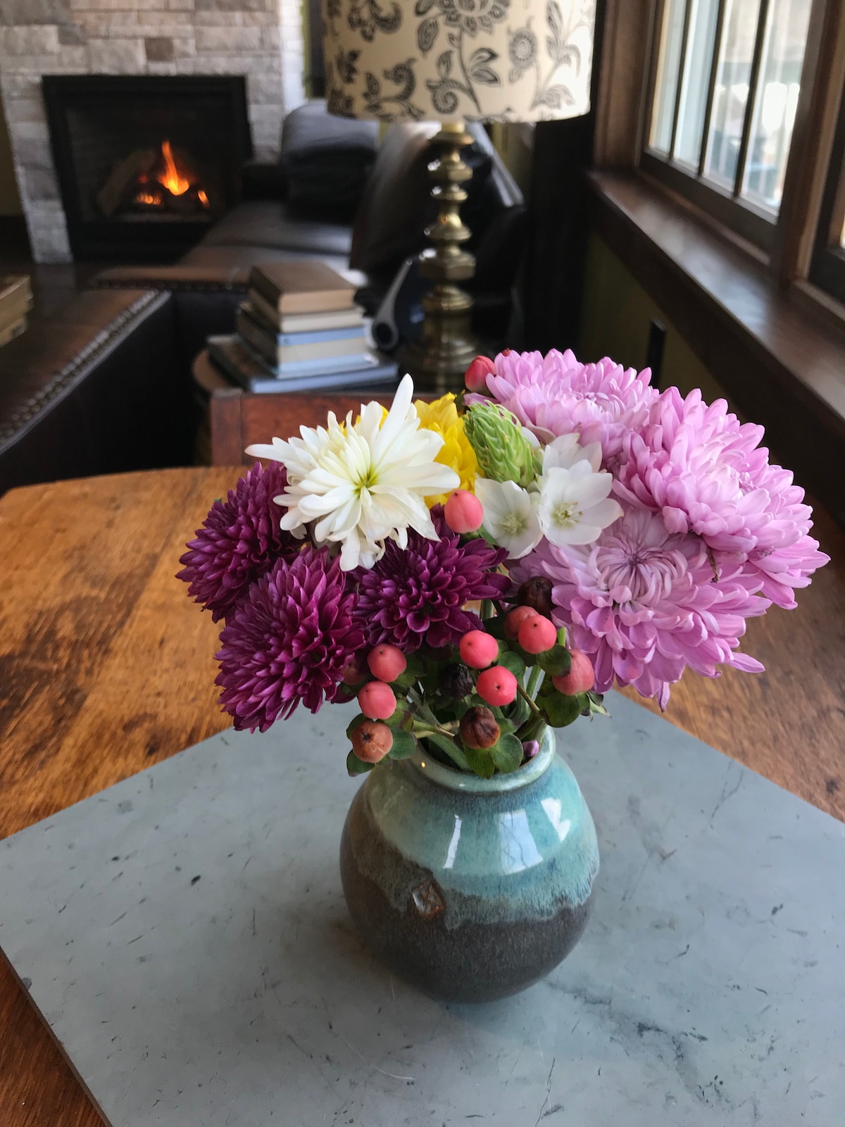 A vase filled with colorful flowers sits on a wooden table. The arrangement includes purple, white, and yellow blooms along with green foliage and small berries. In the background, a cozy gas fireplace is partially visible, adding warmth to the inviting indoor space.