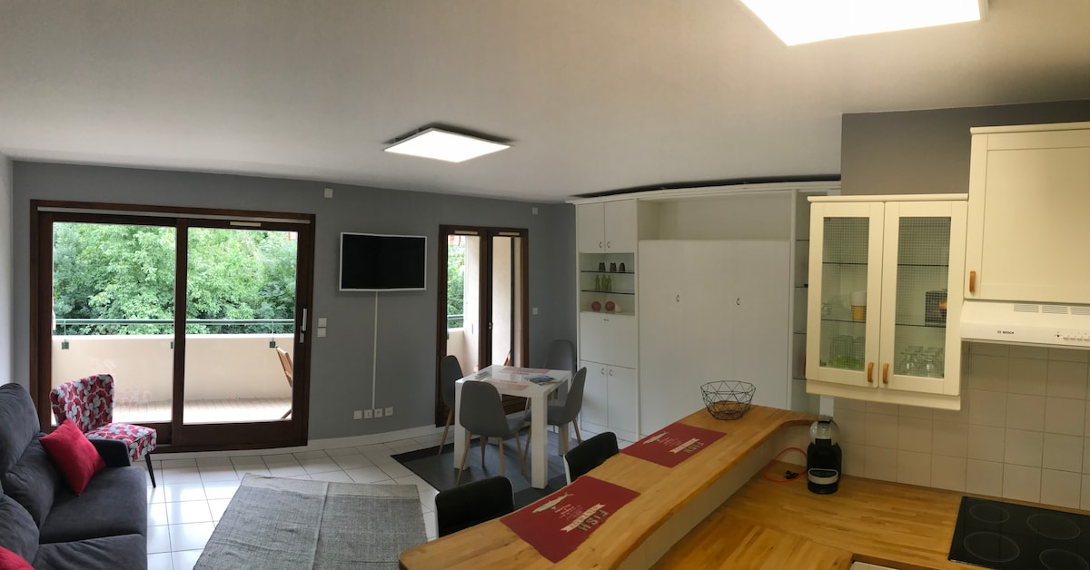A spacious studio interior is highlighted, featuring a well-equipped kitchen with wooden countertops and a dining area. A comfortable seating arrangement includes a gray sofa and a patterned armchair. Large windows frame a view of greenery, allowing natural light to fill the space.