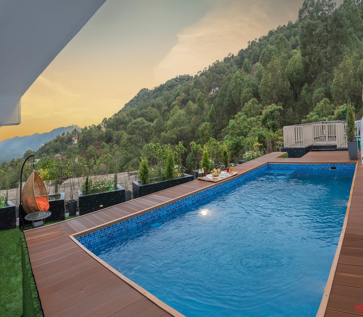 A private swimming pool is surrounded by wooden decking and views of lush greenery. Comfortable seating is provided nearby, along with refreshments. The tranquil setting captures the essence of relaxation, with mountains in the background and a soft glow from the setting sun.