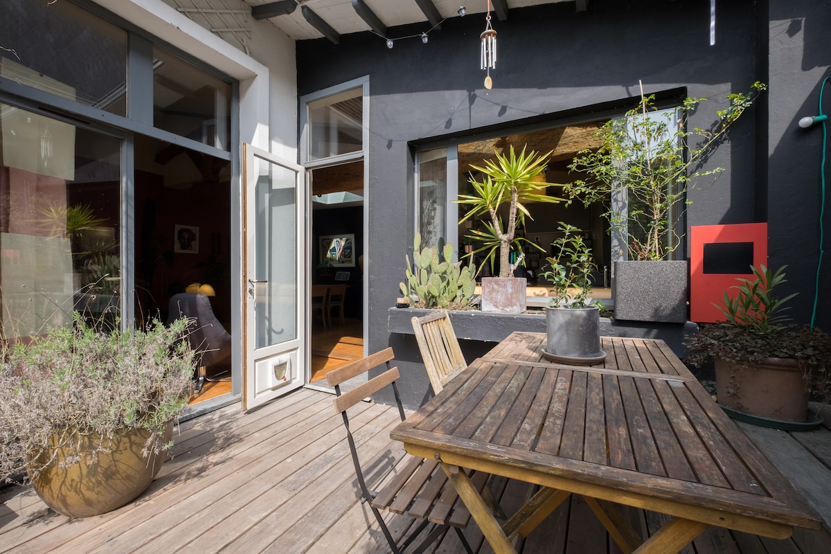 A wooden terrace features a rectangular table surrounded by chairs, with potted plants and a small cactus adding greenery. Large glass doors open to the interior space, creating a seamless transition. Natural sunlight highlights the wooden deck, enhancing the overall ambiance.