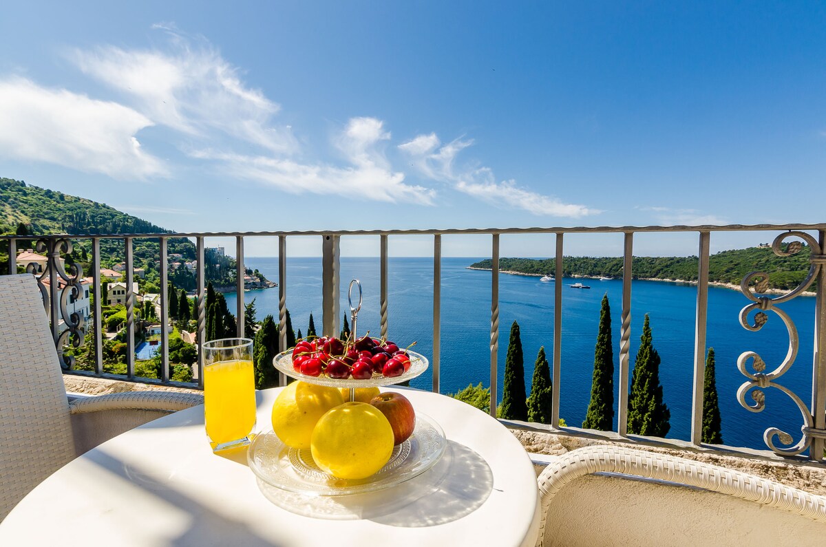 A small table is set with a plate of fresh strawberries and apples, accompanied by a glass of orange juice. The view showcases a serene blue sea under clear skies, framed by lush green hills and tall cypress trees along the coast.