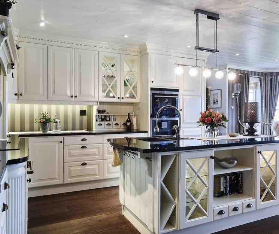 A spacious kitchen features cream cabinetry with glass fronted accents and a large island in the center. Modern appliances, including an oven and dishwasher, are integrated into the design. Soft lighting illuminates the area, complemented by decorative plants and a fresh flower arrangement.