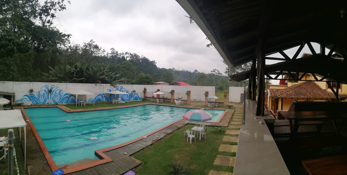 An outdoor swimming pool is surrounded by green lawns and features shaded seating areas. Colorful umbrellas and lounge chairs are arranged around the pool, with a mural of waves painted on the adjacent wall. The backdrop consists of distant mountains under a cloudy sky.