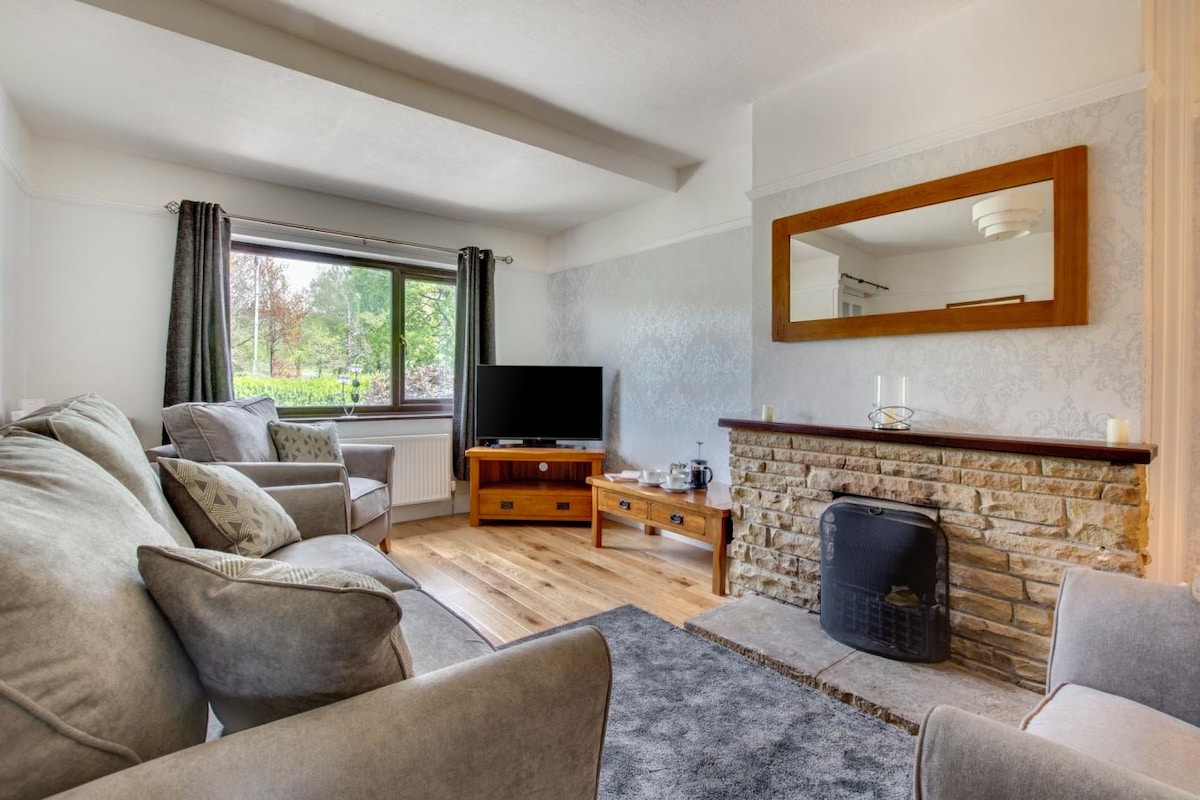 The living area features soft seating arrangements, including a plush gray sofa and armchairs. A flat-screen television is positioned on a wooden console. Natural light enters through a large window, complementing the neutral décor and stone fireplace that adds a touch of warmth to the space.