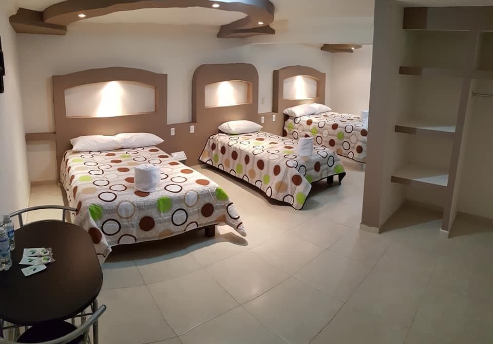 A spacious room contains three beds with patterned bedspreads featuring circles in various colors. A small round table with chairs is positioned in one corner, and shelves line the adjacent wall. Soft lighting highlights the neutral-colored walls of the room.