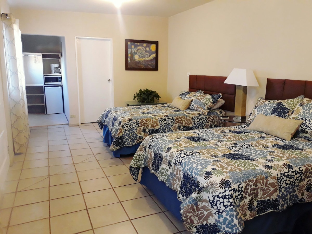 A guest room features two spacious beds dressed in floral patterned bedding, complemented by decorative pillows. A bedside lamp is positioned between the beds. A door leads to a kitchenette area, and a piece of wall art adds character to the neutral-toned walls.