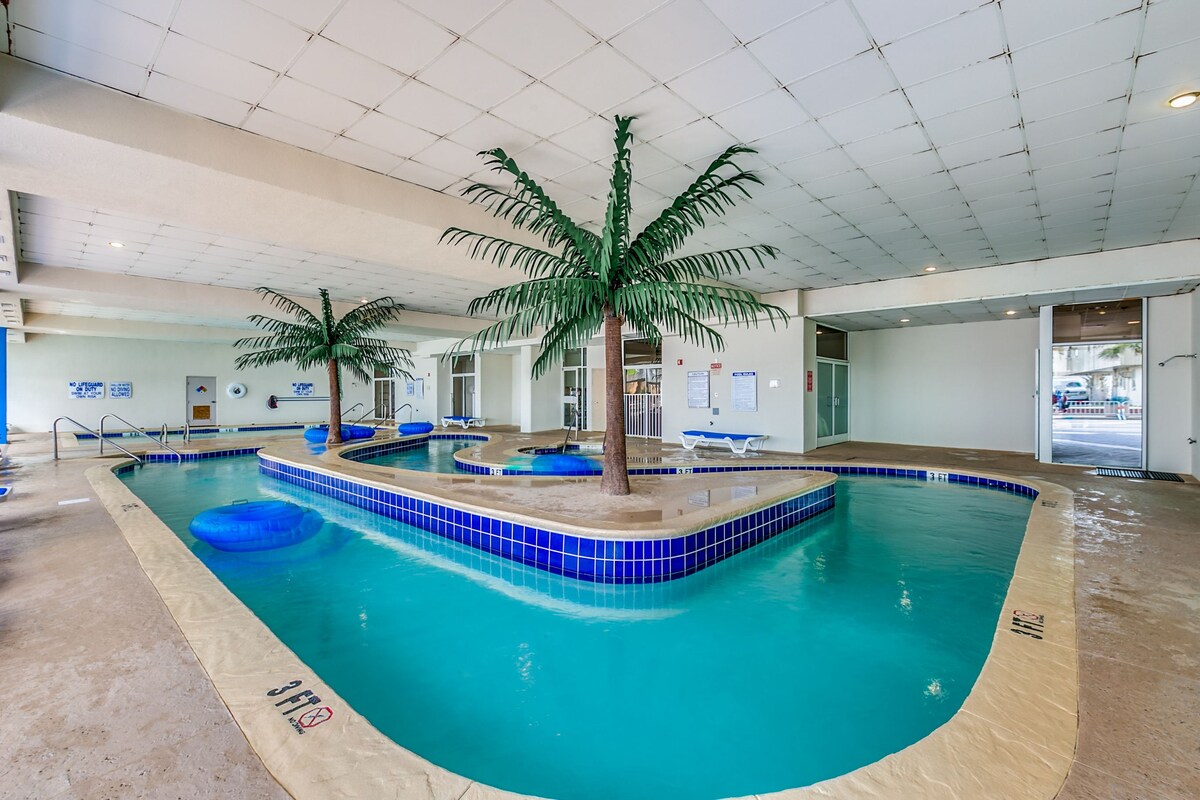An indoor pool area is highlighted by clear turquoise waters and tropical palm tree features. The pool is surrounded by a smooth, beige deck, with blue loungers positioned nearby. Floatation devices are visible in the water, enhancing the inviting atmosphere of the space.