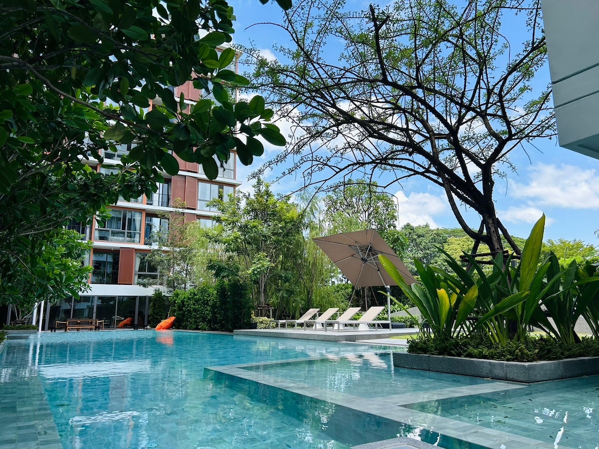 A serene outdoor swimming pool is surrounded by lush greenery and modern lounge chairs. A large umbrella provides shade beside the water. The pool reflects the clear blue sky and is bordered by ornamental plants and trees, creating a peaceful ambiance.