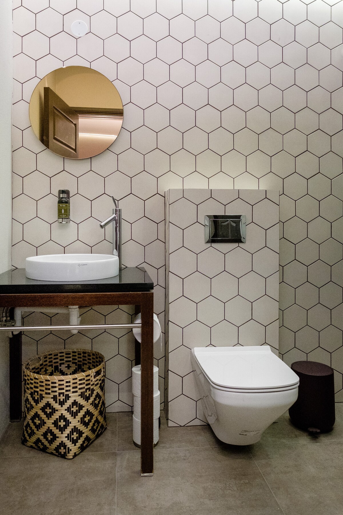 A modern bathroom features a clean design with hexagon-patterned tiles. A round mirror hangs above a circular sink on a dark wood vanity. A white toilet is positioned next to a decorative woven basket, providing a blend of functionality and style.